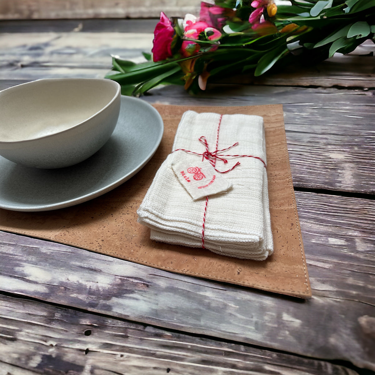 Cork placemat with a gray plate, cream bowl, and tied white napkins on a wooden table beside flowers.
