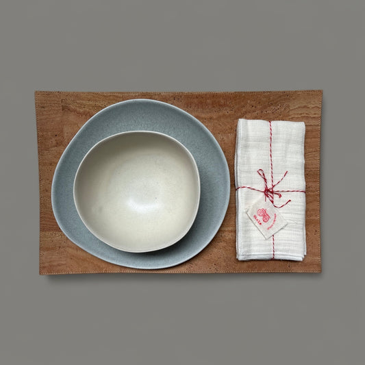 Gray plate and cream bowl beside white napkins tied with red twine on a rectangular cork placemat.