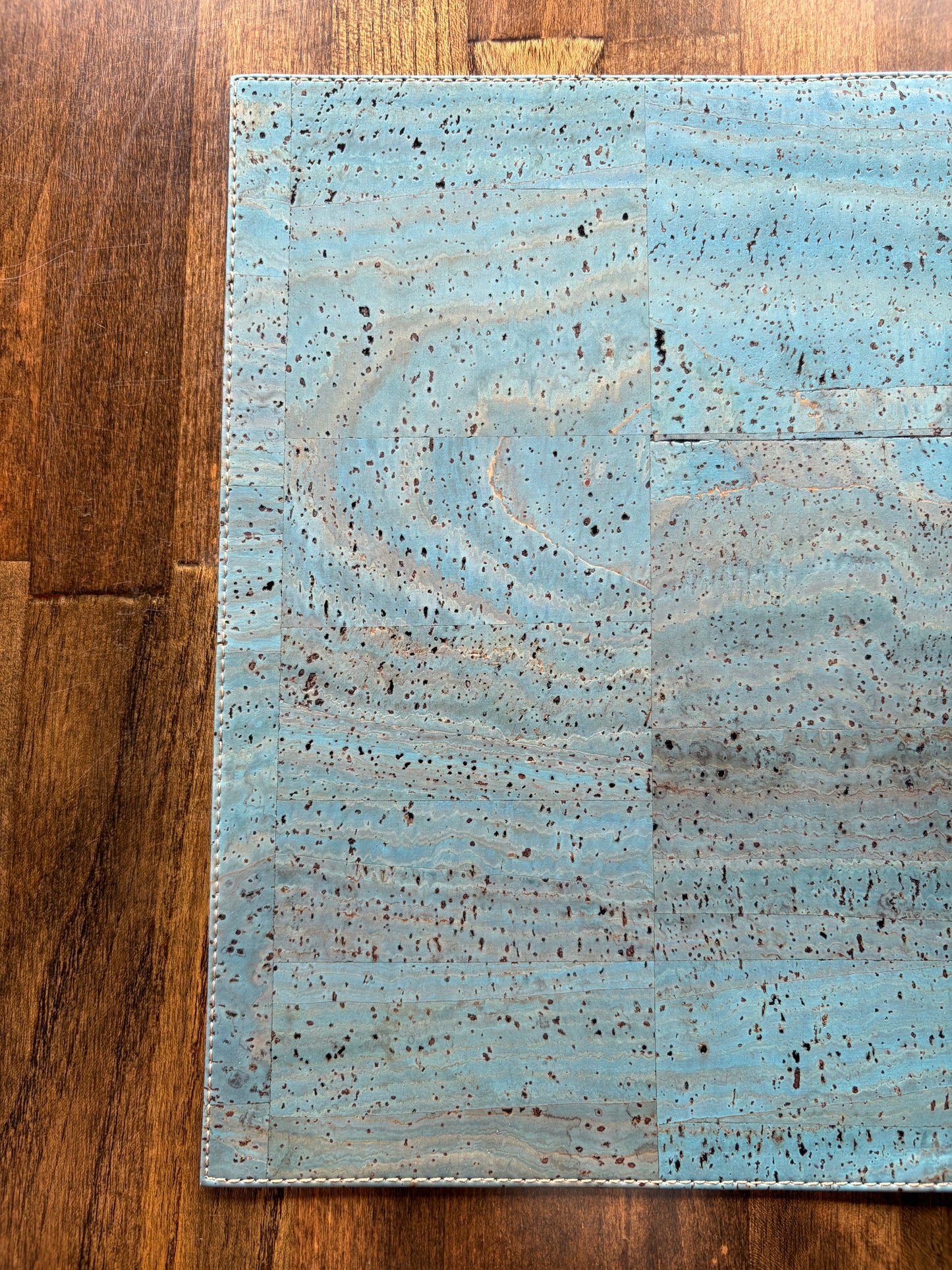 Close-up of light blue cork placemat showing natural cork flecks, subtle swirls, panel joins, and stitched edges on wood.