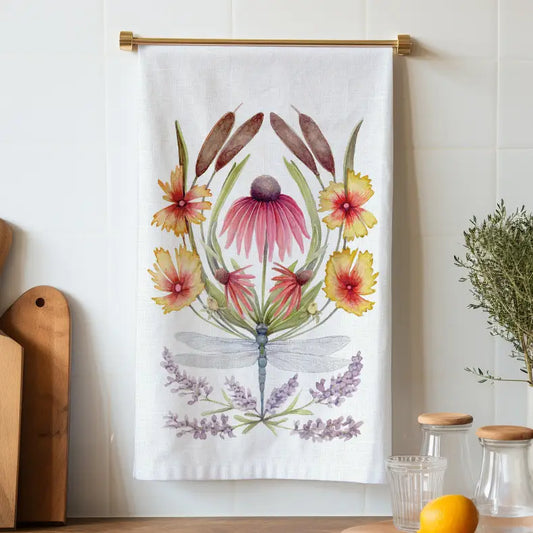 White tea towel hanging from a brass rod, illustrated with a dragonfly, cattails, pink and yellow flowers, and lavender sprigs.