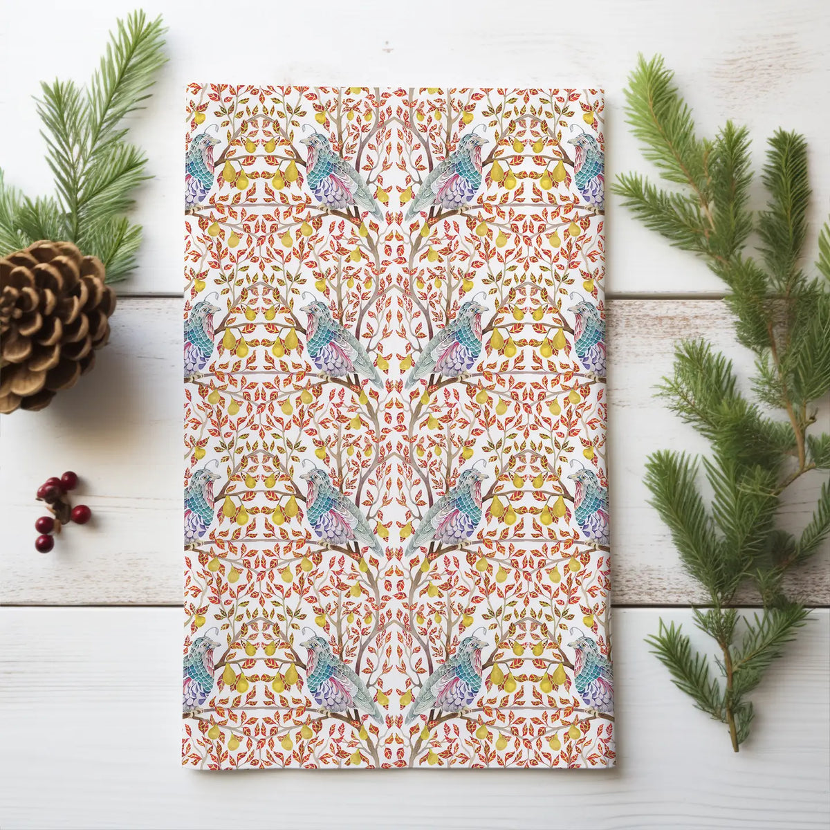 Folded tea towel with blue-green partridges, yellow pears, and red leaves on white wood beside evergreen sprigs and a pinecone.