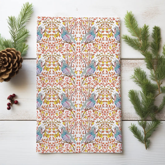 Folded tea towel with blue-green partridges, yellow pears, and red leaves on white wood beside evergreen sprigs and a pinecone.